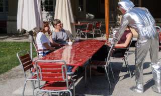 Eine Person in silbernem Anzug stellt einen Stuhl an einen besetzten Tisch.